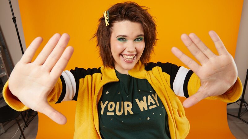 A smiling woman with short, curly hair, wearing a yellow and black jacket and a t-shirt that says "YOUR WAY”.