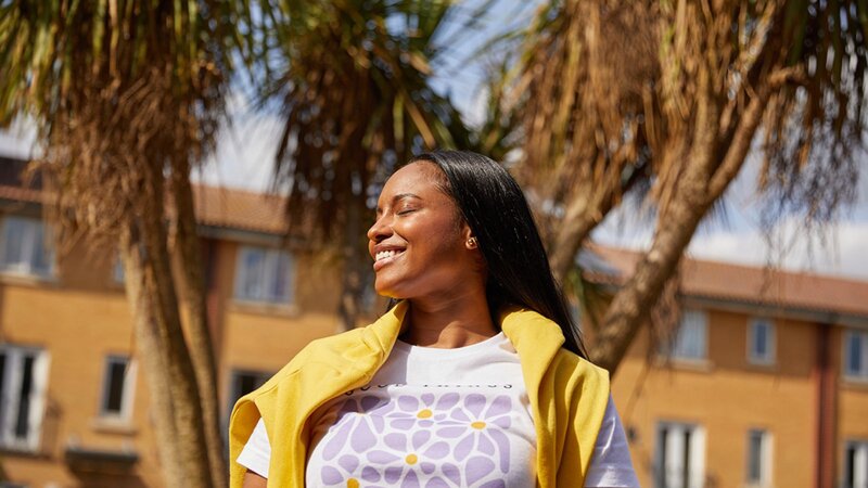 A woman with long hair is smiling while wearing a yellow vest over a white shirt with a floral design, standing outdoors.