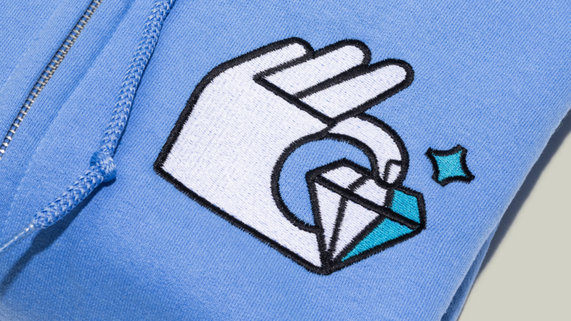 A close-up of a blue hoodie showing an embroidered design of a hand making an "OK" gesture, with a small diamond shape and a star beside it.