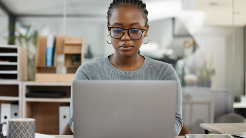 A woman in glasses, wearing a gray sweater, is looking down at a laptop.