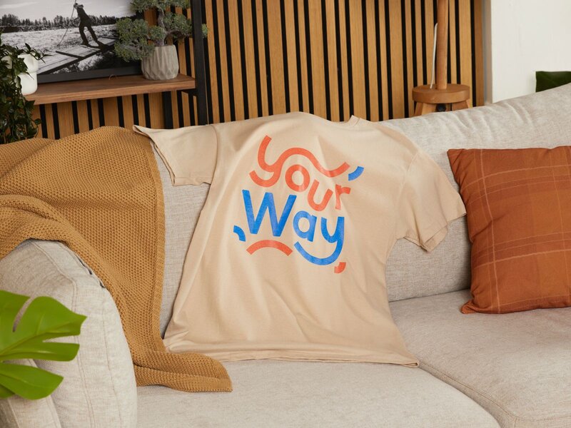 A beige t-shirt with the text "Your Way" in blue and red, placed on a light-colored sofa with a throw blanket and decorative pillows.
