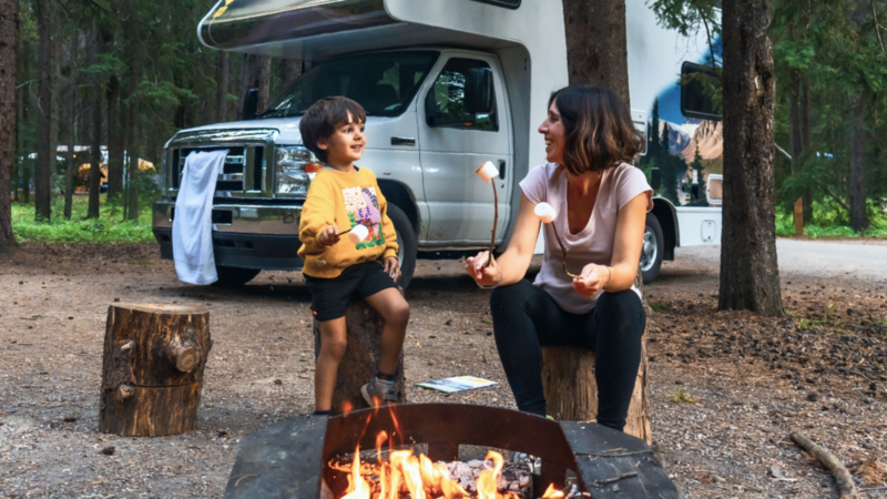 A woman and child toast marshmallows over a campfire in a forest, near an RV.