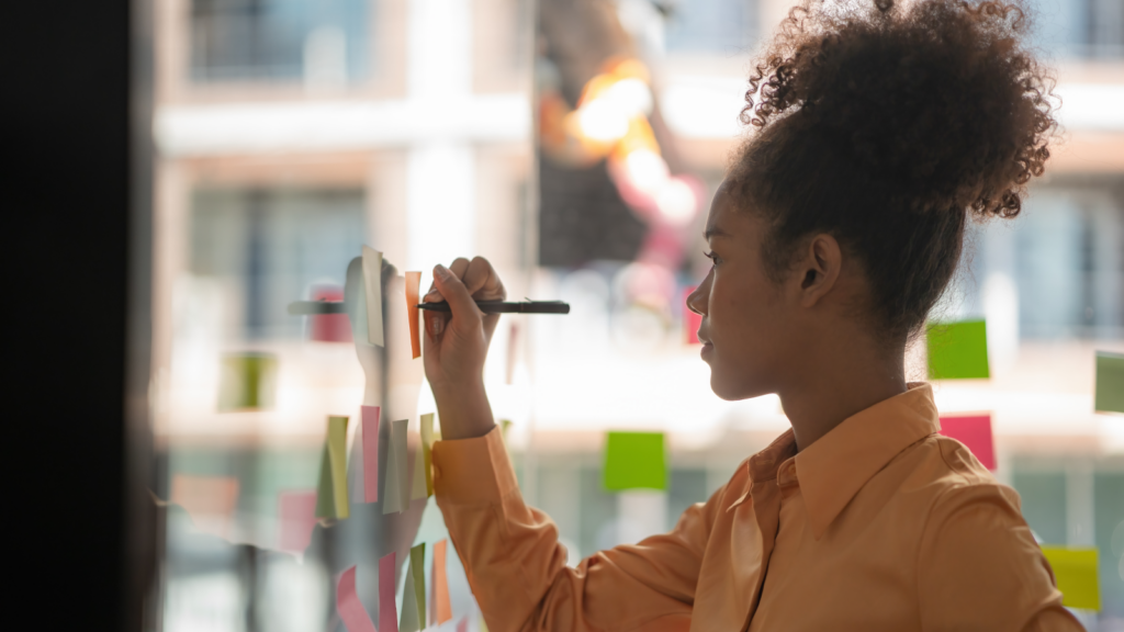 A woman writing on colorful sticky notes affixed to a clear glass wall, focused on organizing her thoughts.