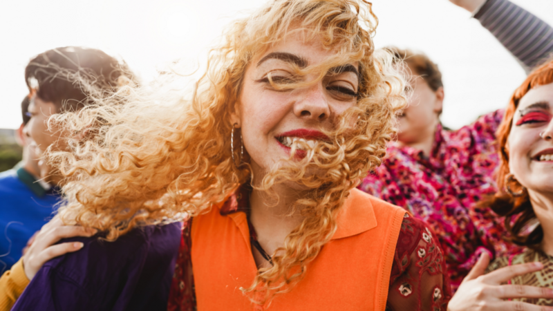 A joyful group of people with a woman in front wearing bright orange, smiling, with windswept curly hair.