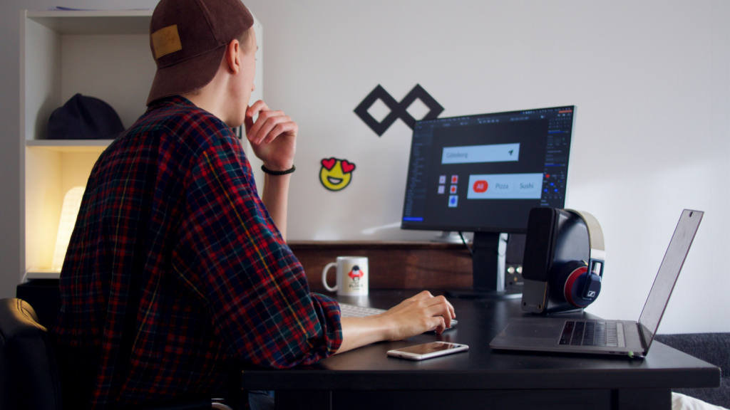 A young man in a checkered shirt, wearing a hat, sitting at a desk, looking at a monitor while doing graphic design work.