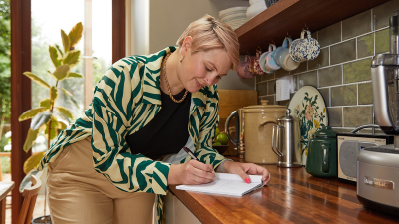 A woman taking notes in a notepad, kitchen in the background.
