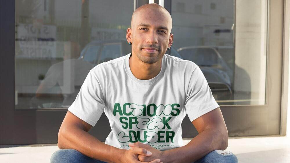A man sitting in front of a door, wearing a white Gildan 2000 t-shirt with a “Actions speak louder” print.