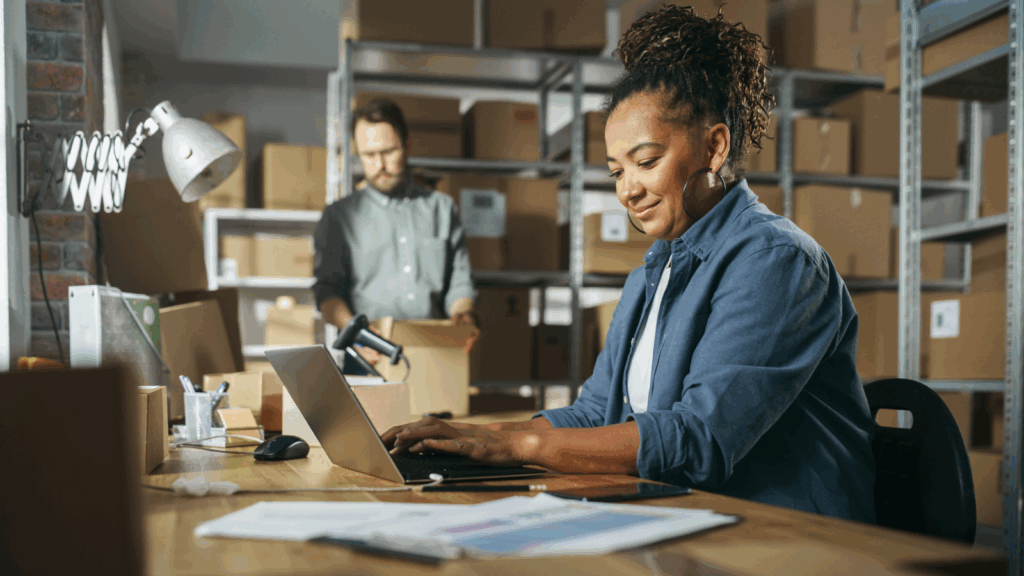 A woman focused on her laptop while seated in a busy warehouse filled with shelves and boxes.