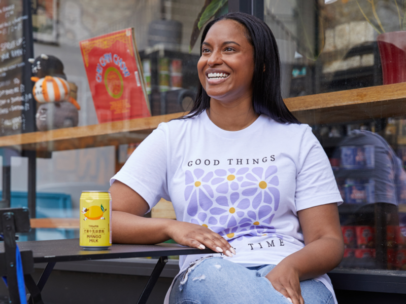 A woman seated at a table, wearing a vibrant print-on-demand t-shirt, exuding a sense of positivity and contentment.