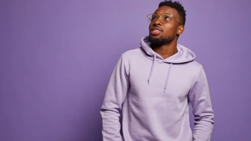 A man in a light-purple hoodie standing in front of a purple background.