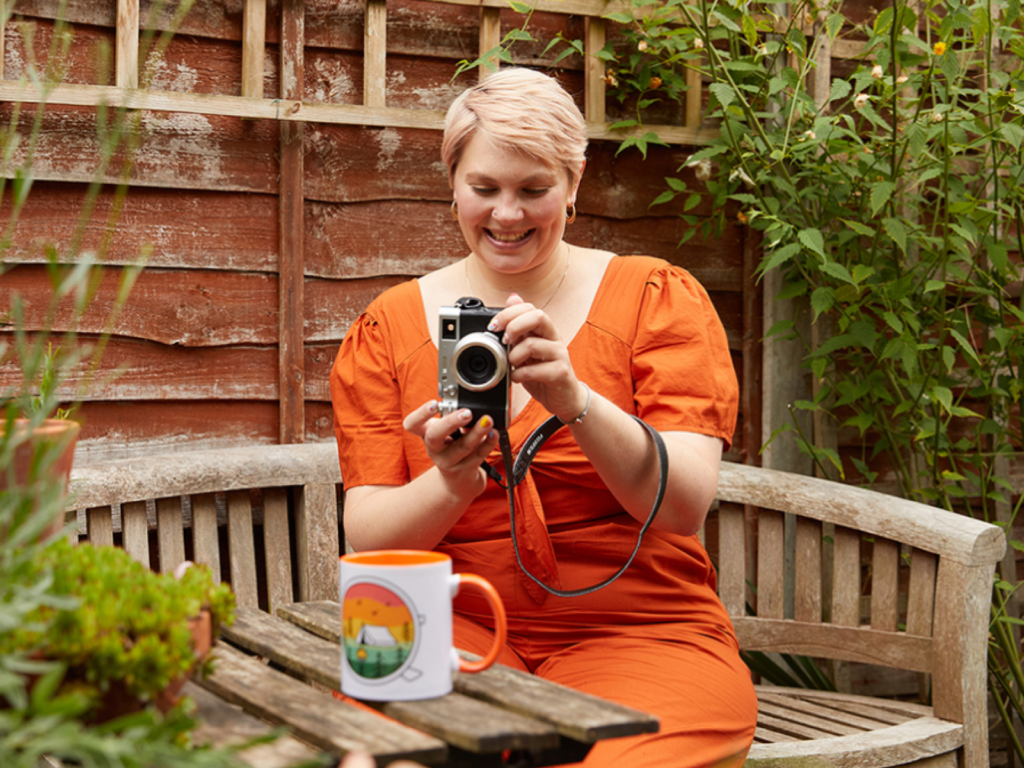 A woman with short hair, wearing an orange dress, takes a picture of a colorful mug in a garden setting.