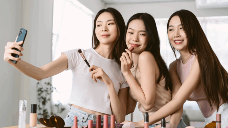 Three women are holding make-up products and recording themselves using a mobile phone.