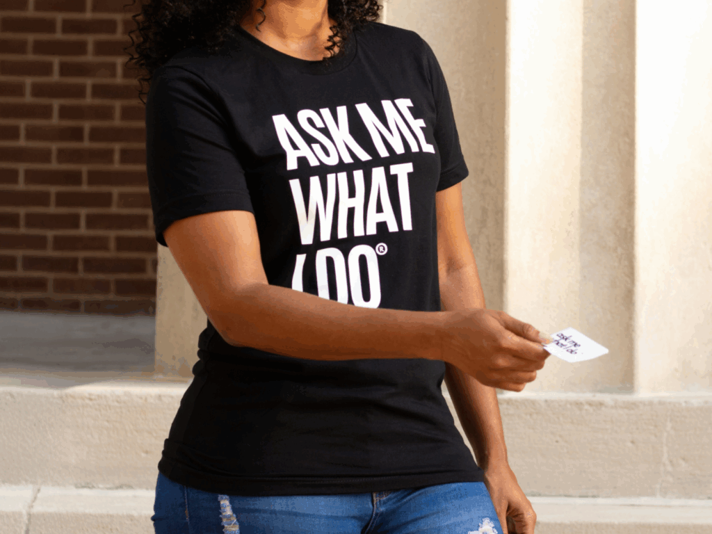 A woman in a black “Ask me what I do” t-shirt is handing out a business card.