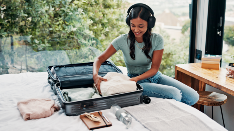 A woman sits on a bed packing a suitcase, wearing headphones and a green shirt. She's folding clothes, with a wooden table nearby.