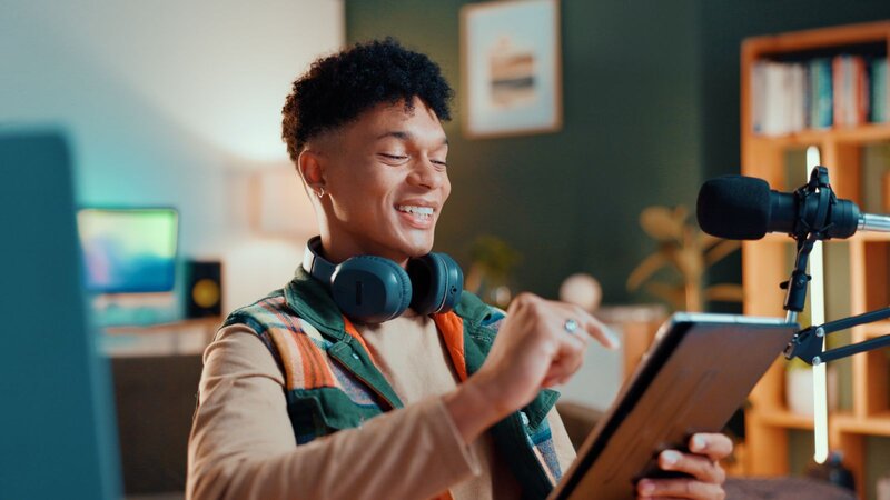 A young person wearing headphones smiles as they use a tablet in a cozy, well-lit room.