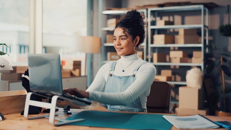 A young woman sitting at a desk, working on a laptop, in a well-lit room with boxes in the background.