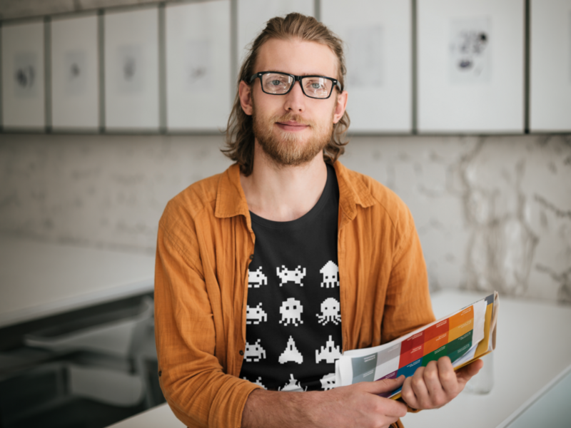 A man with glasses and a beard smiles while holding colorful charts. He wears a black shirt with white space invaders and an orange jacket.