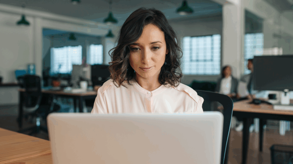 A woman with curly hair is focused on a laptop in a modern office.