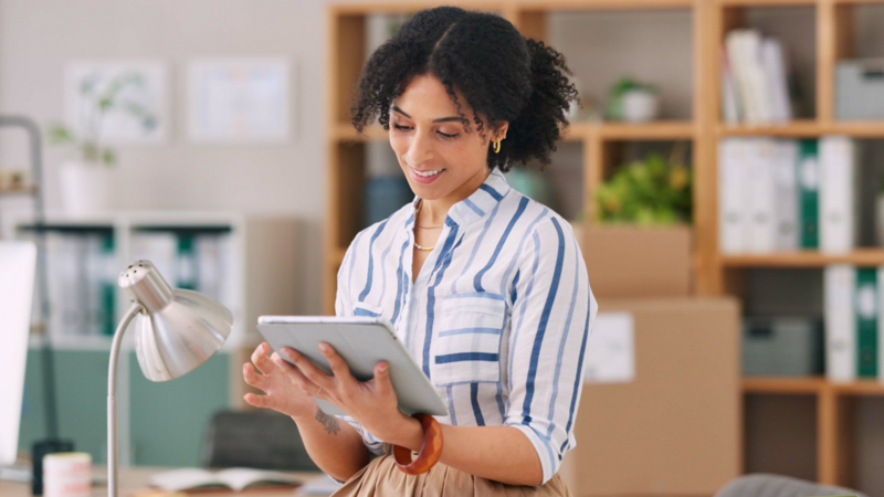 A smiling woman in a striped shirt uses a tablet in an office.