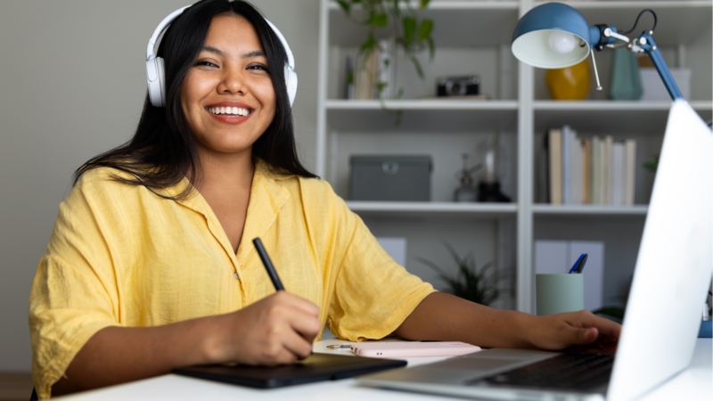 A smiling woman in headphones uses a drawing tablet and laptop.