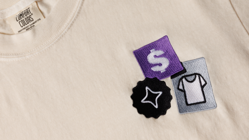 A close-up of a light-colored t-shirt featuring three colorful patches: a purple dollar sign, a gray clothing tag, and a black star-shaped badge.