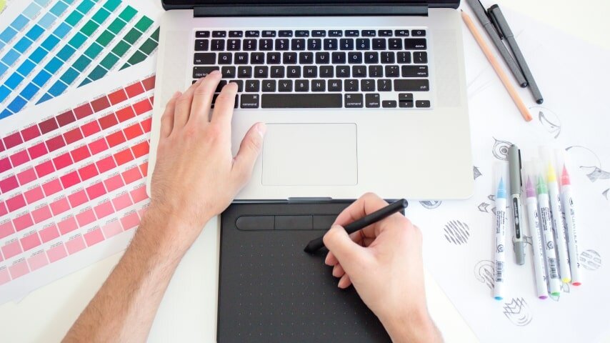 A person drawing something on a tablet in front of a laptop.