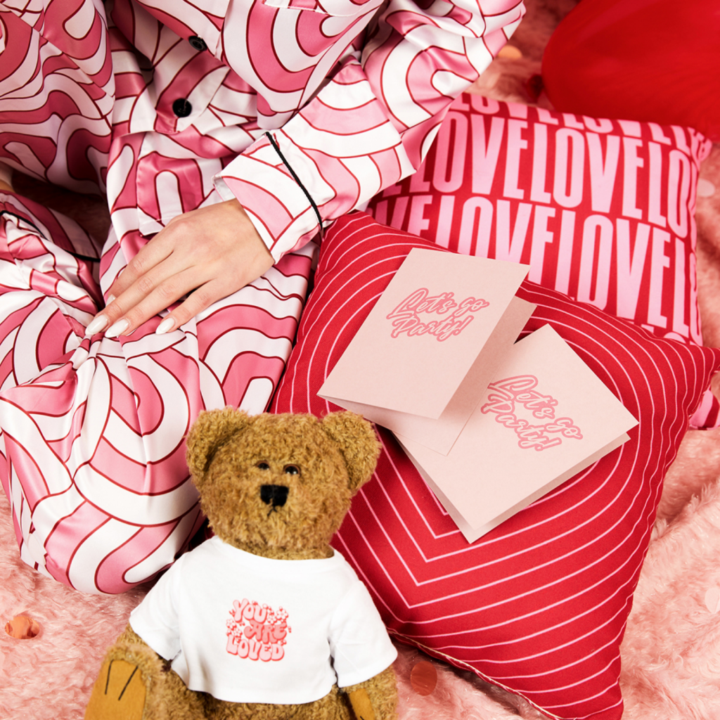 A woman in cute pajamas snuggles a teddy bear, leaning against a fluffy pillow and reading a Valentine’s Day postcard.