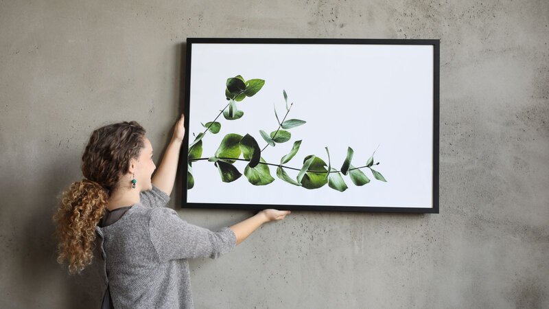 A woman with curly hair holding a framed artwork featuring green leaves against a textured gray wall.