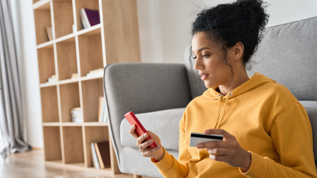 A woman in a yellow hoodie holds a credit card while looking at her phone, appearing focused and engaged in a transaction.