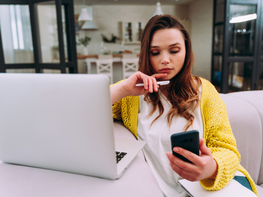A woman sitting in front of a laptop, holding a pen in one hand and a mobile phone in the other.