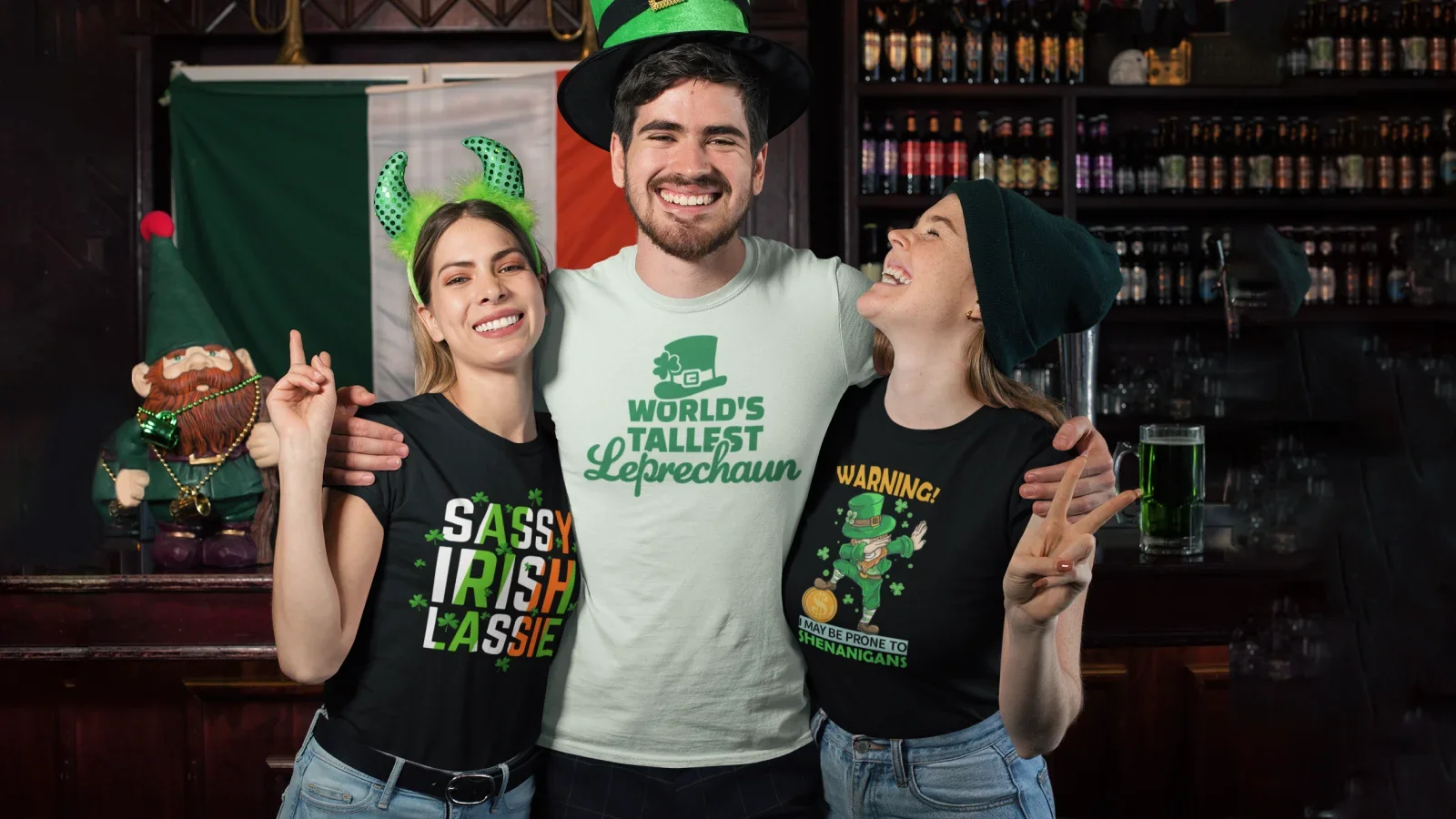 Three friends smiling and posing together for a photo in front of a lively bar wearing custom St. Patrick’s Day shirts.