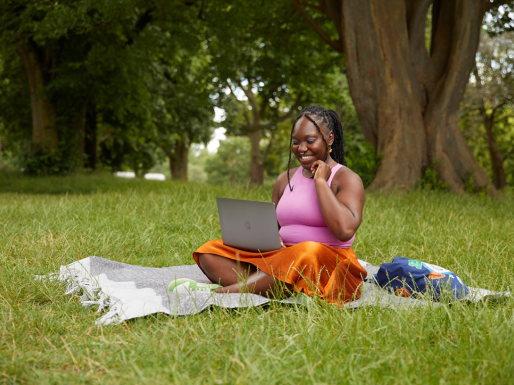 A smiling woman sitting on a blanket in a park and using her laptop to make extra income.
