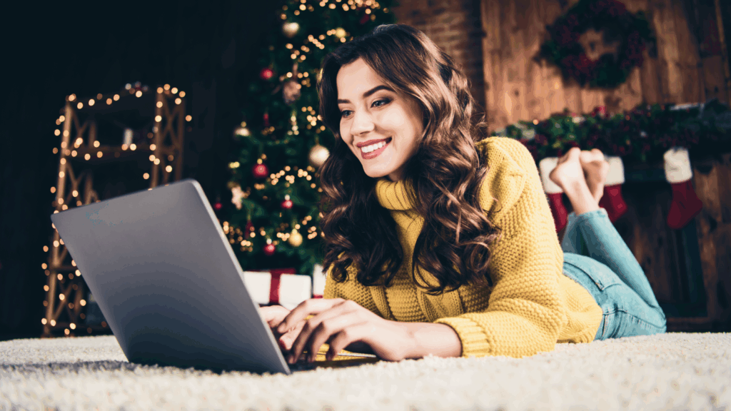 Smiling woman in a yellow sweater using a laptop while lying on a carpet with a decorated Christmas tree and stockings in the background.