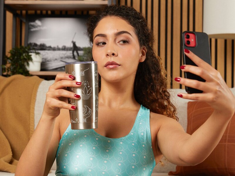 A woman with curly hair takes a selfie while holding a stainless steel tumbler, wearing a light blue tank top, in a cozy indoor setting.