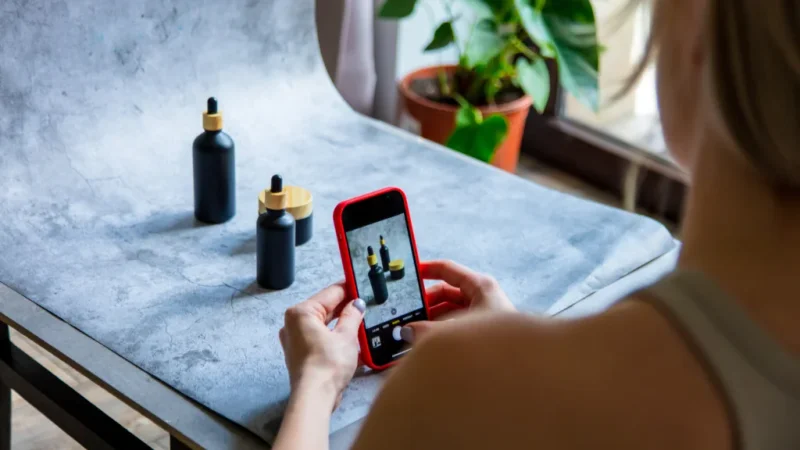 A person photographs three black bottles with wooden lids using a smartphone. The bottles sit on a gray surface near a window.