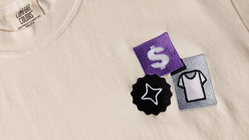 A beige t-shirt with three embroidered patches: a purple square with a dollar sign, a black star-shaped patch, and a grey square with a t-shirt icon.