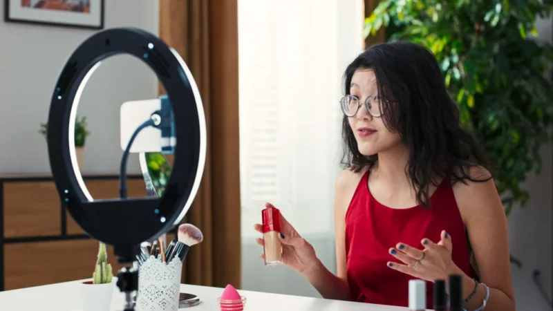 A woman in glasses and a red top is presenting a product while livestreaming using a mobile phone on a tripod with a ring light.
