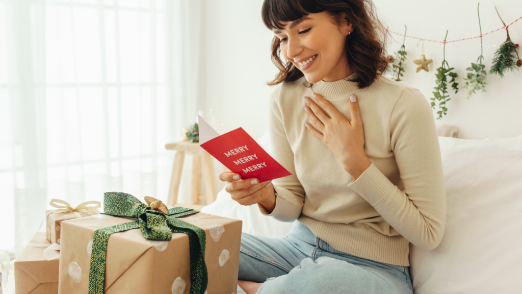 A woman sits on a bed surrounded by presents and a festive postcard, embodying holiday cheer and warmth.
