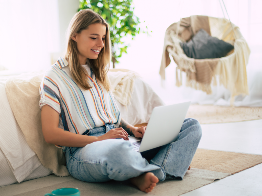 A woman sitting cross-legged on the floor in a cozy bedroom, using her laptop to research how to sell on Etsy and start an art shop.