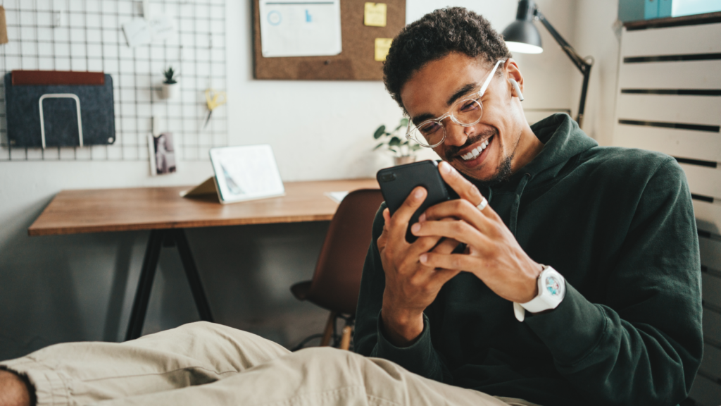 A smiling man with glasses, wearing a green sweater and beige pants, is sitting in an office, legs on the table, looking at his phone.
