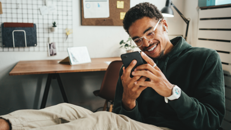 A smiling man with glasses, wearing a green sweater and beige pants, is sitting in an office, legs on the table, looking at his phone.