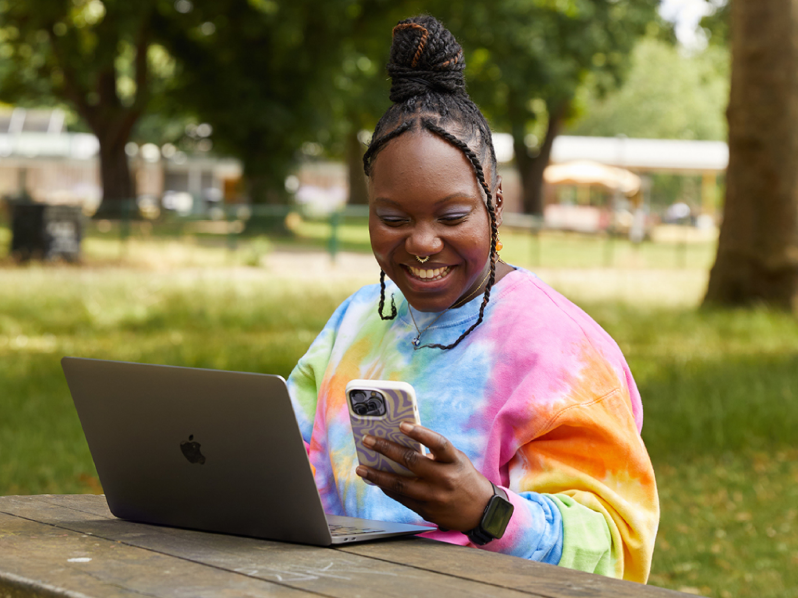 A smiling woman is looking at her phone while sitting at a bench in a park and using a laptop.