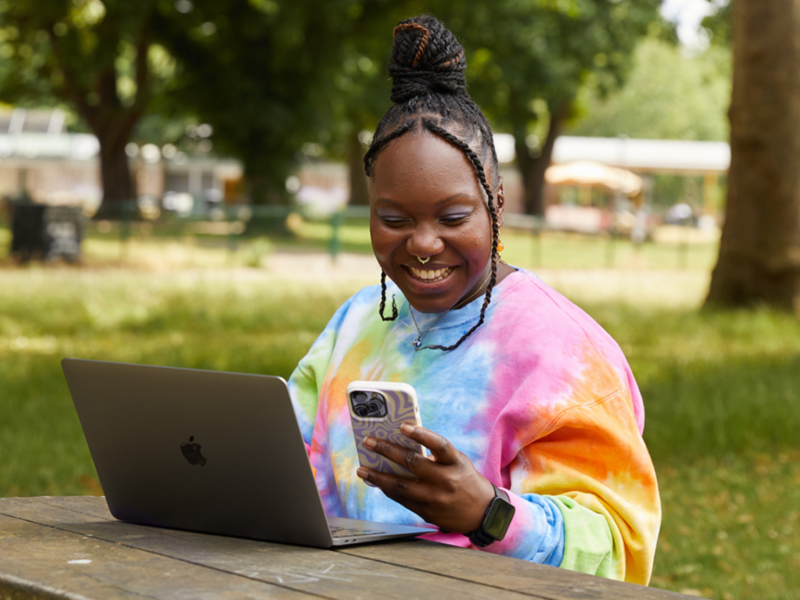 A smiling woman is looking at her phone while sitting at a bench in a park and using a laptop.