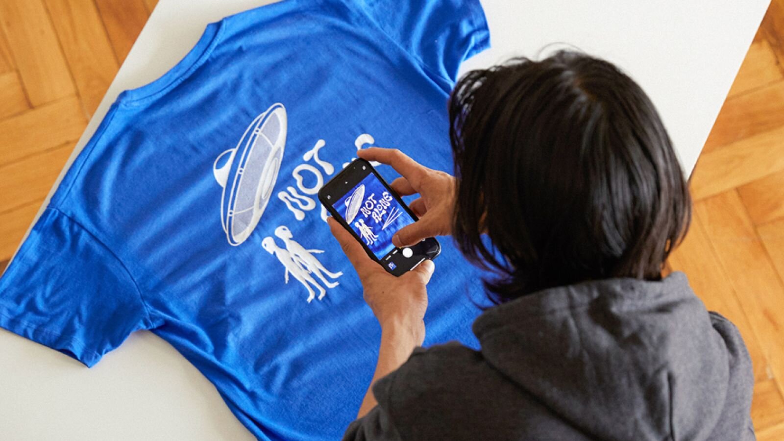 A man in a hoodie photographs a blue t-shirt with a UFO design and the text "NOT ALONE" using a smartphone.