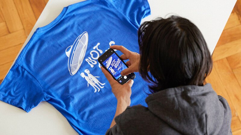 A man in a hoodie photographs a blue t-shirt with a UFO design and the text "NOT ALONE" using a smartphone.
