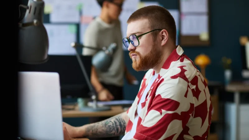 A man with a beard, wearing glasses and a red-and-white floral shirt, is focused on a laptop. A busy office in the background.