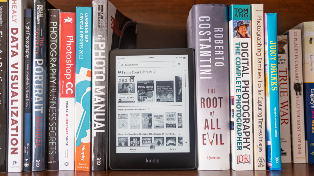 A Kindle rests on a bookshelf beside a neatly stacked collection of various books, showcasing a cozy reading space.