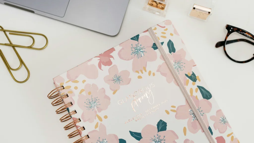 A floral-designed planner in pink next to a notebook with a gold frame, highlighting a chic and organized workspace.