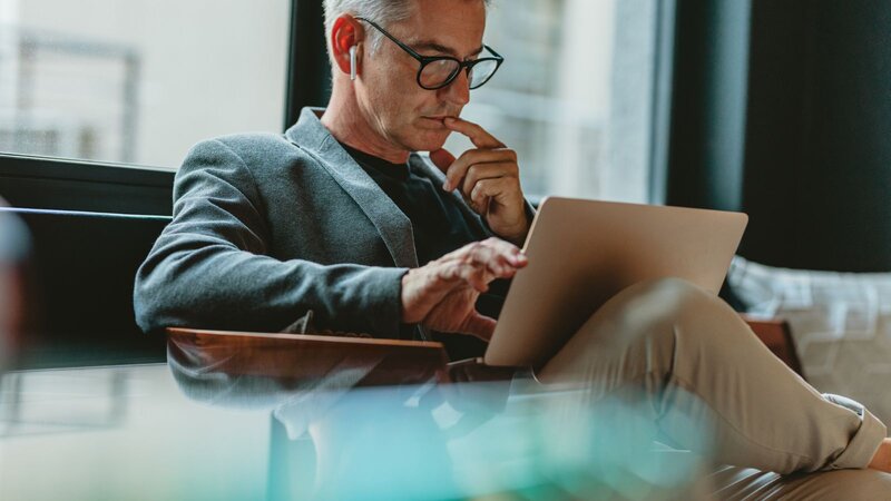 A man in glasses and a blazer sits comfortably while working on a tablet, learning about Etsy ads, holding his chin in thought.