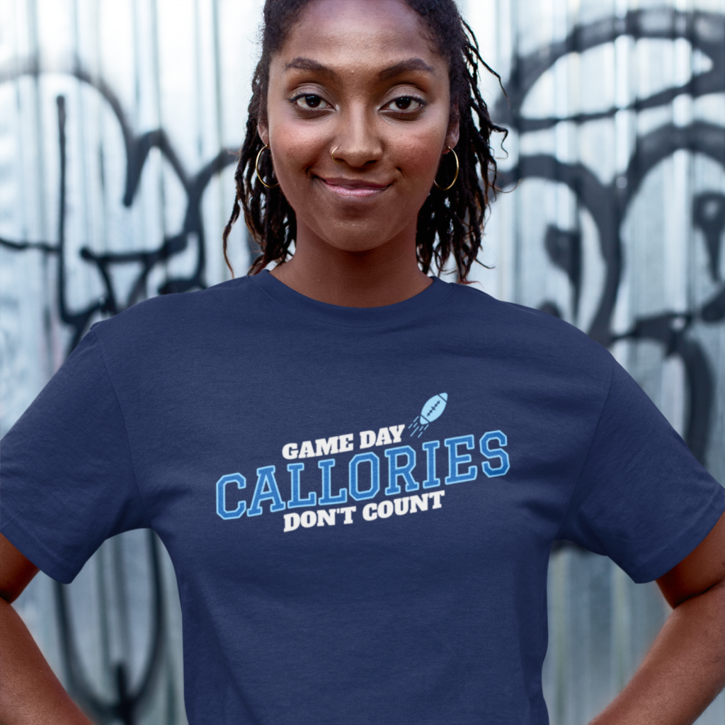 A smiling woman wearing a navy t-shirt with the text "Game Day Calories Don't Count" and a football graphic. Background features graffiti on metal.